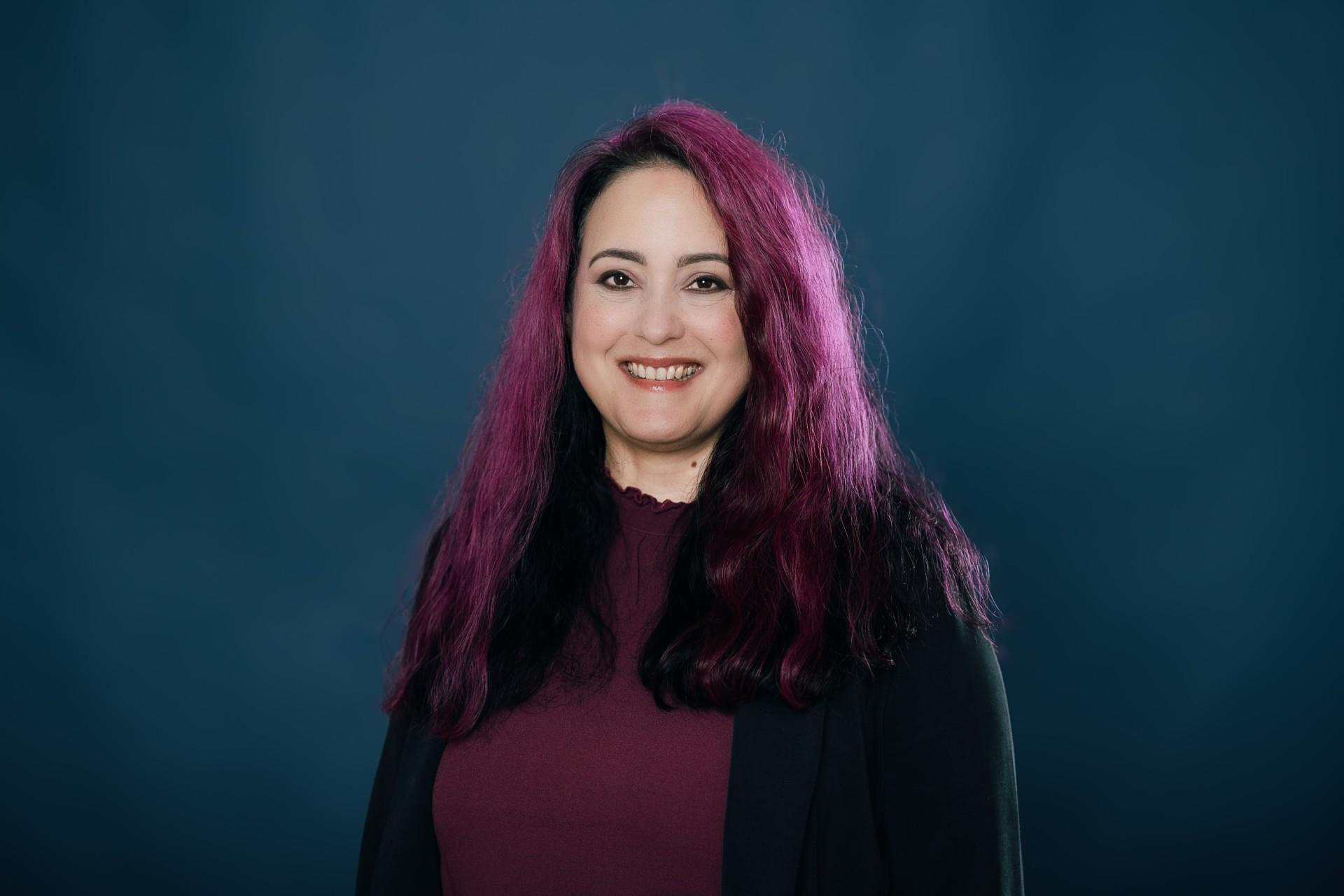 Frau mit violett-schwarzen Haaren, dunklem Oberteil und schwarzer Bluse lächelt in Kamera