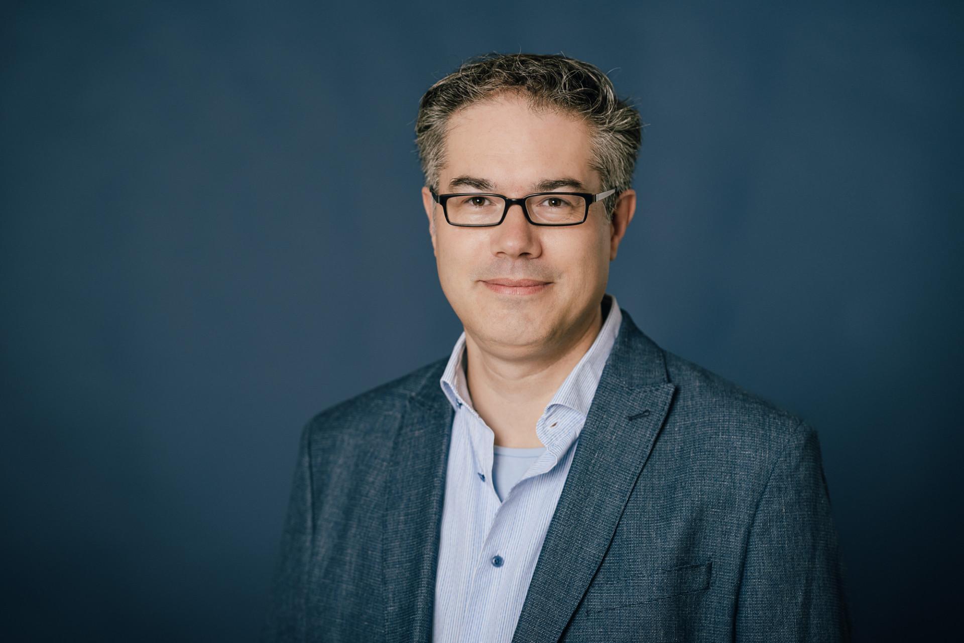 Dr. Sebastian Heer | Transformationsforschung Mann mit Brille und kurzen lockigen Haaren und Hemd vor blauem Hintergrund